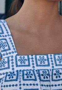 Biała tkanina z niebieskimi haftami kwiatowymi. Kwadratowy dekolt i szerokie ramiączka. Wzór składa się z kwadratów, okręgów i motywów kwiatowych.