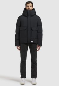 Zwarte pufferjack met een capuchon, voorzien van twee borstzakken, een hoge kraag en elastische manchetten. Draag over donkere jeans en witte sneakers.