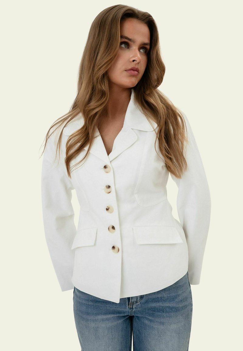 Femme aux longs cheveux ondulés portant un blazer blanc ajusté à cinq boutons et poches avant, assorti à un jean bleu.