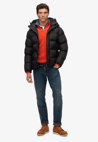 Sort jakke med hætte, orange sweater, blåternet skjorte, blå jeans og brune læderstøvler. Inkluderer sølvur på håndleddet.