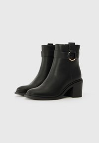 Stivali alla caviglia in pelle nera con punta tonda, ampia fascia e dettagli con anello dorato, e tacco largo. Texture liscia e design minimalista.