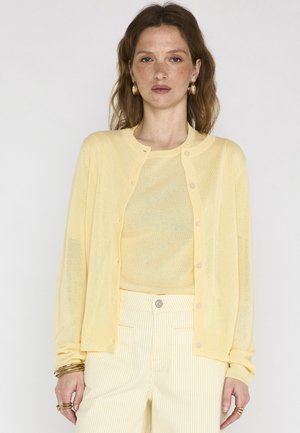 Femme aux cheveux châtain clair portant un cardigan jaune boutonné sur un haut assorti et un pantalon à rayures jaune pâle, accessoirisée de bijoux en or.