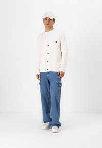 Kremstripete cardigan med treskinnknapper, en lomme, kombinert med blå cargo-jeans og hvite sneakers. Også iført en lys caps.