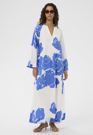 Nő, aki hosszú fehér ruhát visel nagy kék virágmintákkal, kerek napszemüveget és barna, sarokkal ellátott szandált, egyszínű háttér előtt áll.