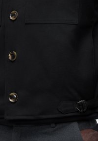 Μαύρο σακάκι με κουμπιά από φελλό και ρυθμιζόμενο πλαϊνό τοκά, φορεμένο πάνω από σκούρο γκρι παντελόνι.