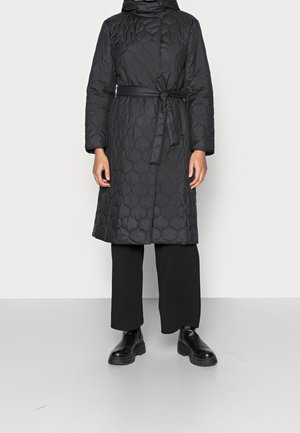 Manteau long matelassé noir avec ceinture nouée à la taille, porté avec un pantalon large noir et des bottes épaisses noires, sur fond clair uni.