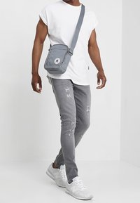 Bolso mensajero gris hecho de un tejido duradero, con un parche de logotipo en la parte frontal, correa ajustable y cierre con cremallera. El modelo lleva una camiseta blanca y jeans grises.