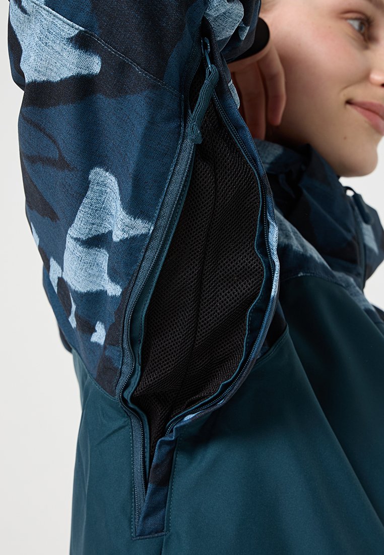 Blau gemusterte Jacke mit einem netzgefütterten Einsatz unter dem Arm, die ein schlankes, funktionales Design und kontrastierendes dunkel-türkisfarbenes Material aufweist.