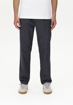 Pantalones oscuros de color gris, hechos de tela texturizada, con un diseño de pierna recta, combinados con zapatillas deportivas de color gris claro y una camiseta blanca lisa.