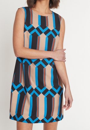 Mouwloze jurk met geometrisch patroon in blauw, zwart, bruin en beige, gedragen door een persoon die één arm over het lichaam kruist.