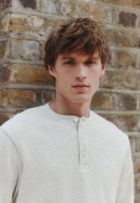 Vaaleanharmaa waffle-kuvioinen pitkähiha-paita, jossa on ribbineulos ja pyöreä pääntie. Kauluksella kolme nappia. Taustalla tiiliseinä.