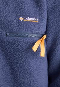 Mornarska flis jakna z žepom na zadrgo in oranžnim trakom ter logotipom podjetja Columbia Sportswear na prsih.