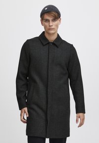 Mørk grå ullfrakk med knappeåpning, spiss krage, sidelommer og teksturert finish. Stylet med en grå strikket lue.