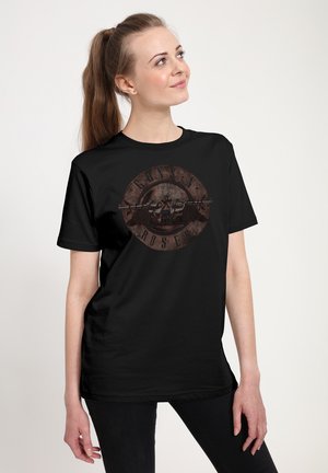 Młoda kobieta z długim kucykiem, ubrana w czarną koszulkę Guns N' Roses, patrzy w górę i na bok na jednolitym tle.