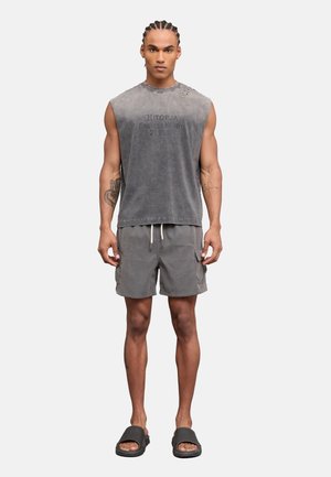 Hombre con cabello trenzado, vestido con una camiseta gris sin mangas con texto, pantalones cortos grises con bolsillos y sandalias negras tipo slide, de pie sobre un fondo blanco.