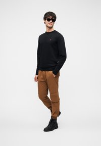 Μαύρο φούτερ με κόκκινο λογότυπο, καφέ παντελόνι cargo με πλαϊνές τσέπες και μαύρα μποτάκια με κορδόνια, στέκεται μπροστά σε ουδέτερο φόντο.