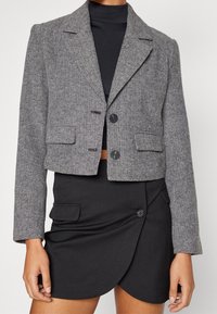 Blazer gris à chevrons avec un design court, deux poches avant et deux boutons, associé à une jupe portefeuille noire avec des boutons.