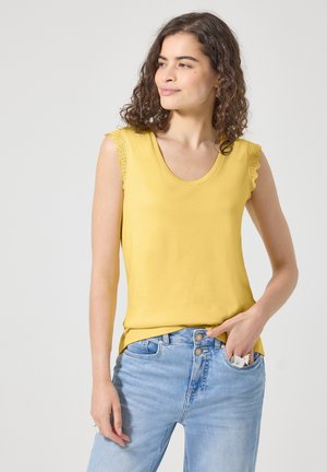 Jonge vrouw met krullend haar die een geel mouwloos topje en lichtblauwe spijkerbroek draagt, met een wit voorwerp in haar zak.