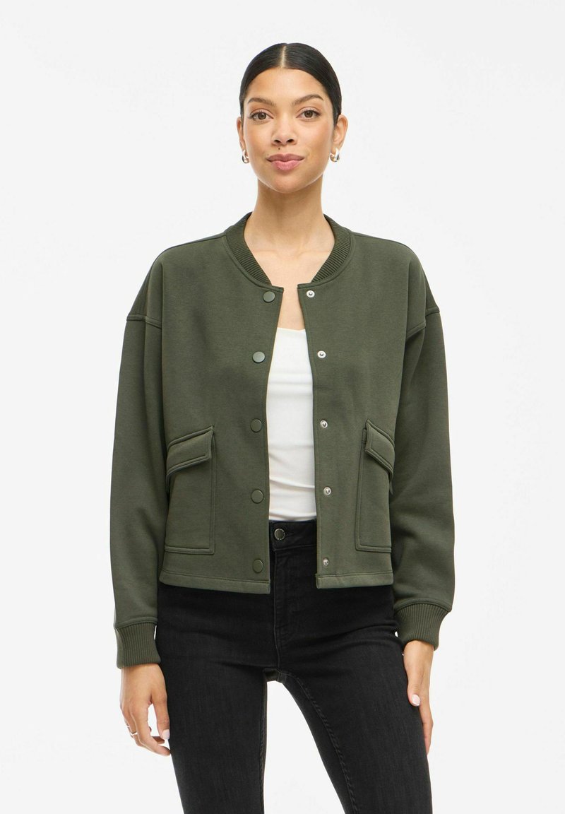 Īss zaļš bumbieru tipa jaka ar apaļu kakla izgriezumu, spiedpogu aizdari, divām priekšējām kabatām, ribotām aprocēm un brīvu piegriezumu. Valkāta virs baltas blūzes.