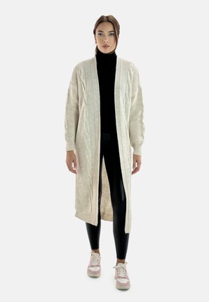 Strickjacke in hellbeige mit Zopfmustern, offener Front, gerippten Kanten, getragen über einem schwarzen Rollkragenpullover und schwarzen Leggings mit pinken Sneakers.