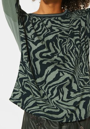 Femme portant une chemise à manches longues avec un imprimé zèbre abstrait vert et noir et un pantalon en cuir noir avec un détail fermeture éclair à la taille.