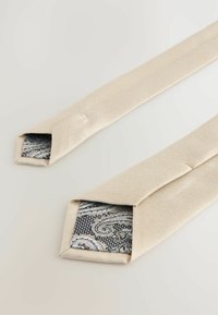 Beige strukturiere Krawatte mit einer Innenseite aus Seide im Paisleymuster. Die Enden sind schräg und zeigen ein dunkleres Blau und ein silbernes Design, das in der Faltung sichtbar ist.