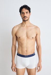 Weiße Boxershorts aus Baumwolle mit einem marineblauen elastischen Bund, der das "GANT"-Logo trägt. Das Modell steht frontal, ohne weitere Kleidung.