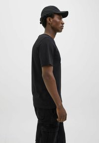 Sort T-shirt med korte ærmer, synlige sømdetaljer og en afslappet pasform, parret med mørke cargo-bukser og en sort kasket.