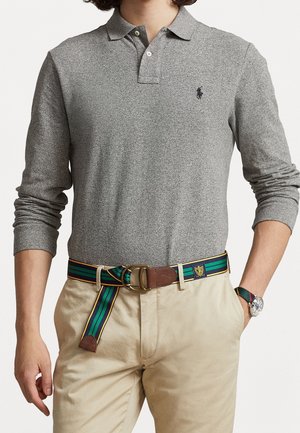 Mężczyzna w szarej koszulce polo z długim rękawem, beżowych spodniach, zielono-niebieskim pasku w paski z elementami skórzanymi oraz w zegarku na nadgarstek z dopasowanym paskiem.