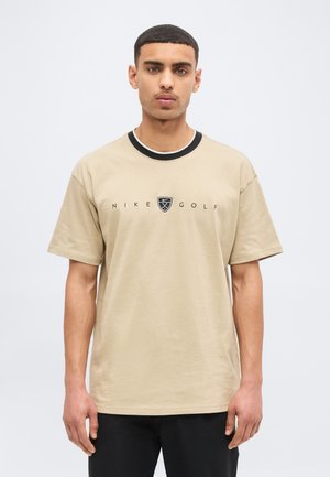 Νεαρός άνδρας που κοιτάει μπροστά, φοράει μπεζ μπλούζα Nike Golf με μαύρη λεπτομέρεια στο γιακά και μαύρο παντελόνι, μπροστά σε απλό φωτεινό φόντο.