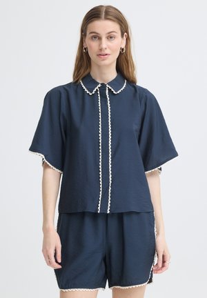 Kvinde iført navyblå top med korte ærmer og matchende shorts med hvid bølget kant på kraven, foran, ærmer og kant.