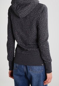 Persona che indossa un hoodie grigio scuro con piccole pois bianchi e rosa, abbinato a jeans in denim blu, mostrato di spalle.