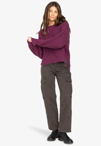 Lila gestrickter Pullover mit lockeren Ärmeln und gerippter Struktur. Kombiniert mit dunklen Cargo-Hosen, die über Seitenstaschen und gestickte Details verfügen.