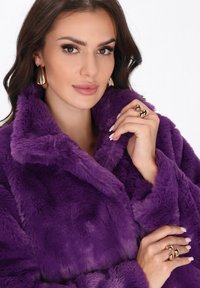 Donna con capelli scuri, che indossa un cappotto di pelliccia sintetica viola, orecchini d'oro e più anelli d'oro, mentre tiene il colletto del cappotto con unghie curate.
