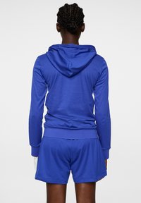 Blå idrottströja med känguruficka och ribbade ärmar, tillsammans med matchande shorts. Materialet verkar slätt och elastiskt.