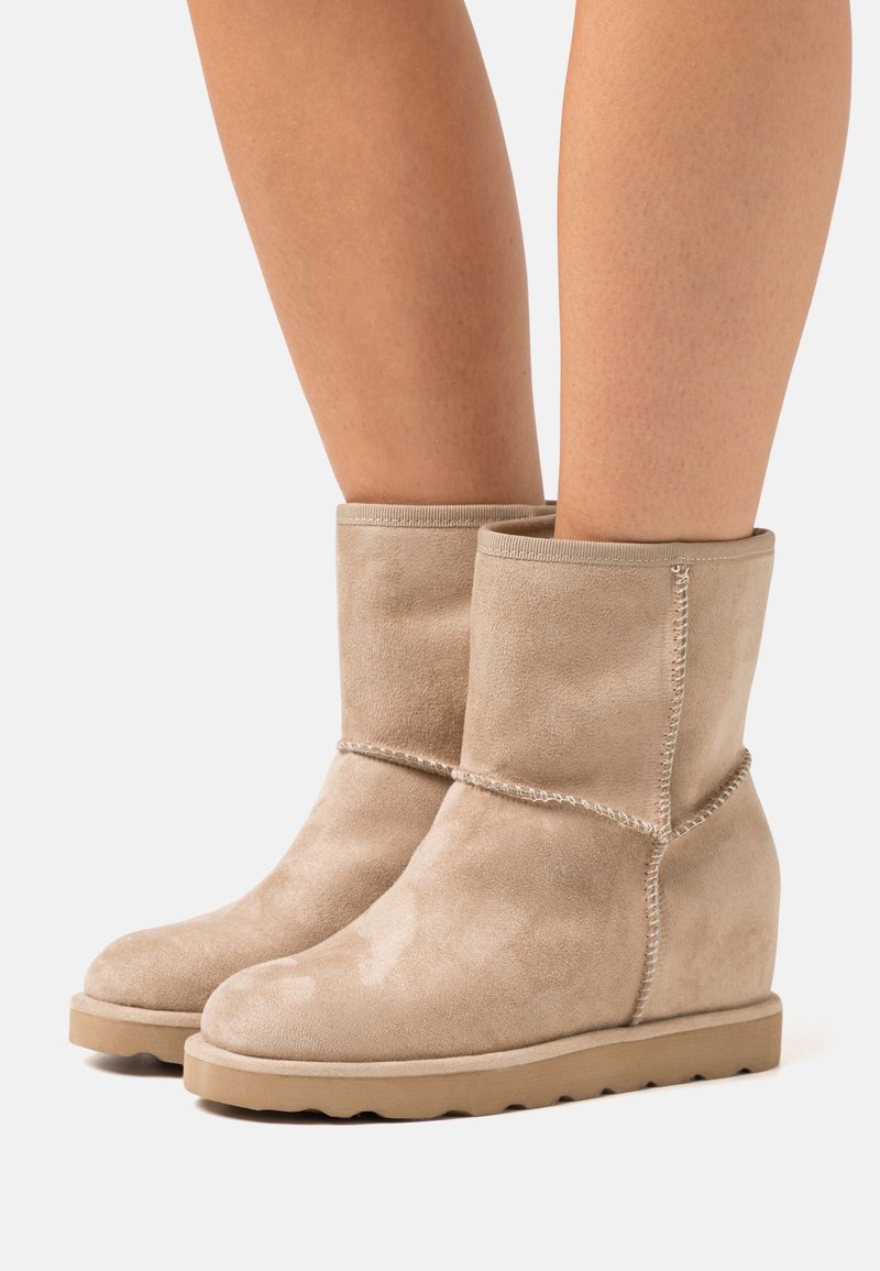 Bottes compensées en daim beige mi-mollet avec coutures apparentes, portées sur des jambes nues sur un fond blanc uni.