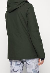 Giacca impermeabile verde con cappuccio, caratterizzata da una texture liscia, design minimalista e orlo regolabile. Indossata sopra pantaloni grigi a motivo camouflage.