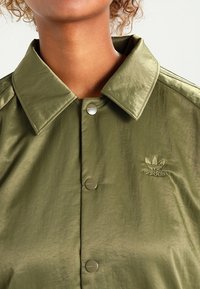 Olivově zelená košile s knoflíky z lesklého materiálu s límcem, zdobená stříbrnými knoflíky a malým vyšívaným logem Adidas.