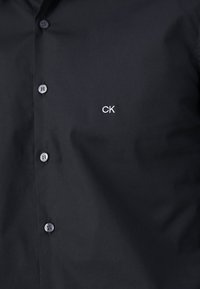 Musta nappipaita, jossa on rintamuksessa kirjailtu "CK"-logo ja näkyvät tummat napit etupaneelissa.