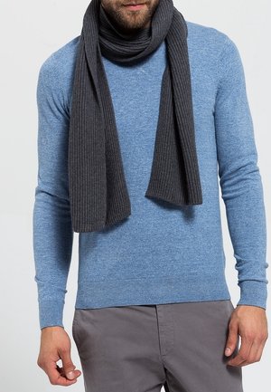Homme portant un pull bleu clair, une écharpe côtelée gris foncé et un pantalon gris, debout les mains détendues le long du corps sur un fond uni.