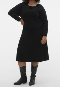 Robe noire tricotée à manches longues, décolleté rond et ourlet évasé. Associée à des bottes de cowboy noires arrivant jusqu'aux genoux.