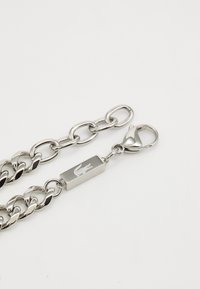 Sølvkjede med store sammenkoblede lenker, som har et rektangulært hardware-stykke med gravert logo og en krabbe-lås for feste.