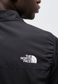 Κοντινό πλάνο ενός ατόμου που φοράει μαύρο μπουφάν North Face, με το λογότυπο στην άνω πλάτη να φαίνεται πάνω σε ουδέτερο φόντο.