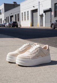 Baskets à plateforme blanches avec des détails en toile et en cuir placées sur un trottoir urbain avec des bâtiments et des voitures garées en arrière-plan.