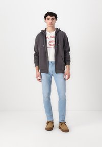 Grå hettejakke med glidelås og snorer, hvit T-skjorte med rødt trykk, lyseblå jeans og brune semskede sko. Enkel, uformell antrekk.