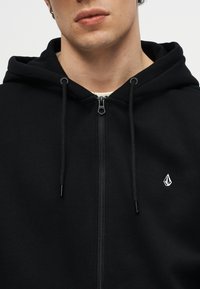 Jeune personne portant un sweat à capuche noir zippé avec des cordons de serrage et un petit logo géométrique blanc sur la poitrine.