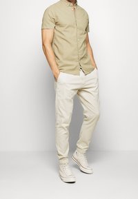 Camisa de oliva de manga corta con cierre de botones, combinada con pantalones tapered color beige claro y zapatillas blancas de caña alta, todo presentado sobre un fondo claro.