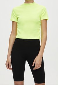 Kobieta w krótkim, neonowożółtym topie i czarnych, wysokich szortach kolarskich na jednolitym tle.