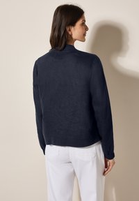 Marinblå linnejacka med en kortad design, räfflad krage och samlade ärmslut, ihopparad med vita byxor. Slät textur.