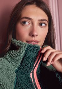 Fliisist jakk kõrge kraega, rohe-mustast tekstuuriga mustriga, roosa tõmblukuga ja pehme ning soe sisemus.